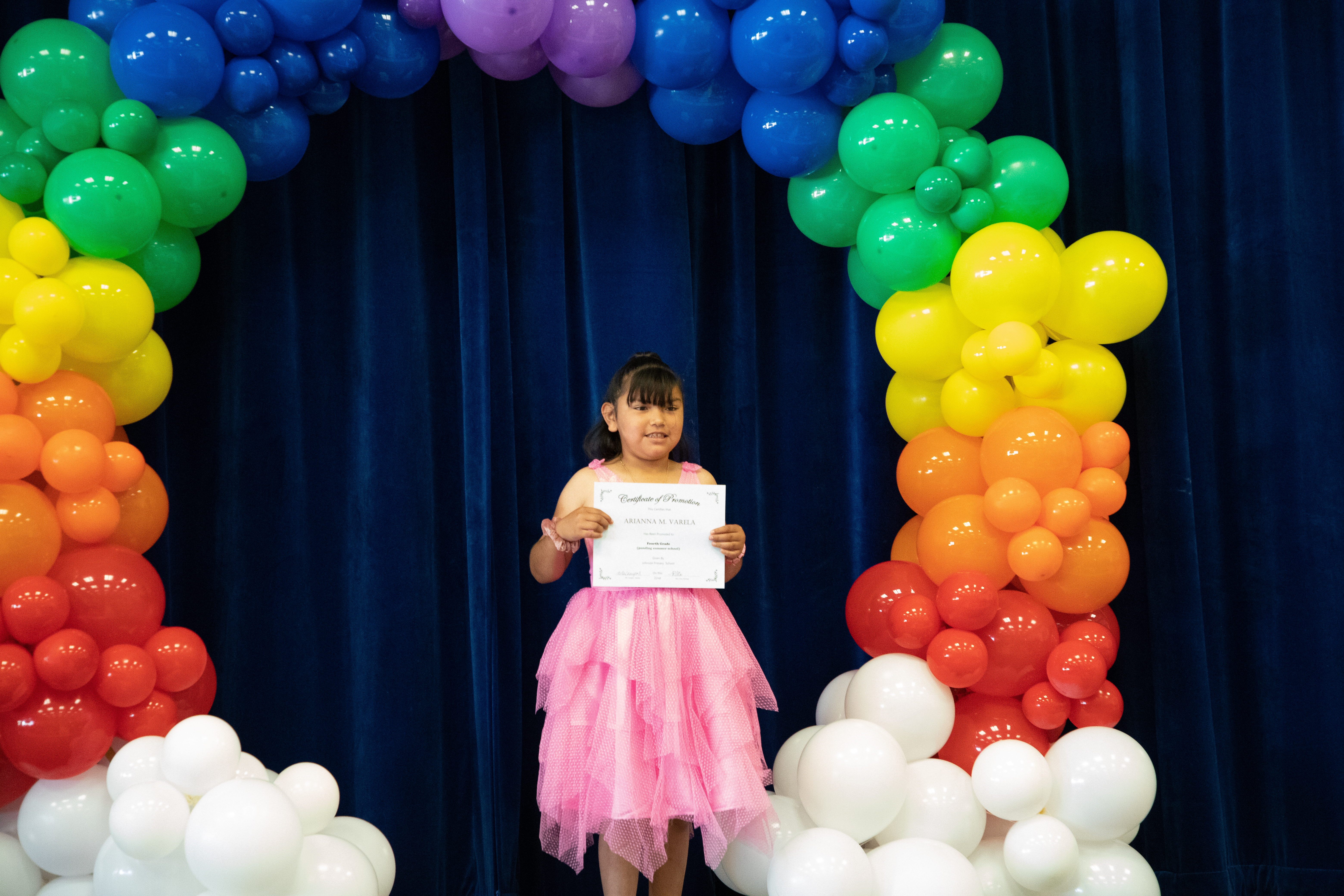 A little girl in a pink dress smiles with her promotion certificate under a rainbow arch of balloons