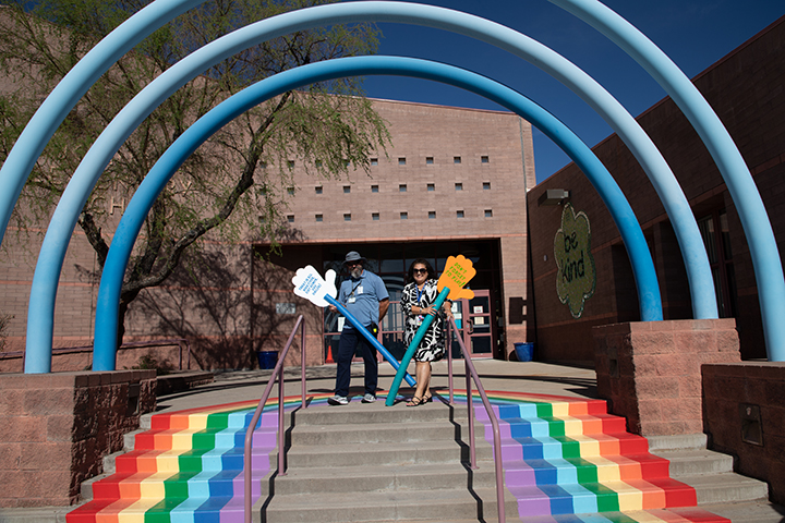 A man and woman stand on top of a rainbow staircase under blue arches, holding up giant hand cutouts on pool noodles