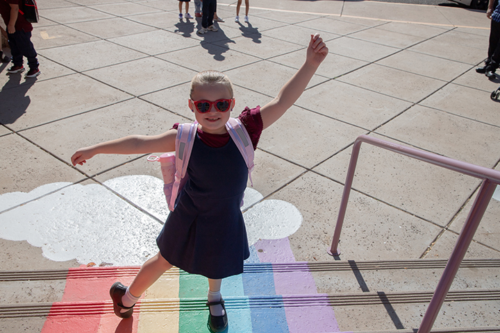 A little girl in a dress and sunglasses holds up an arm as she stands on the rainbow steps