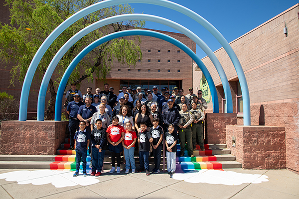 A group of students and first responders stand under the blue arches outside the school