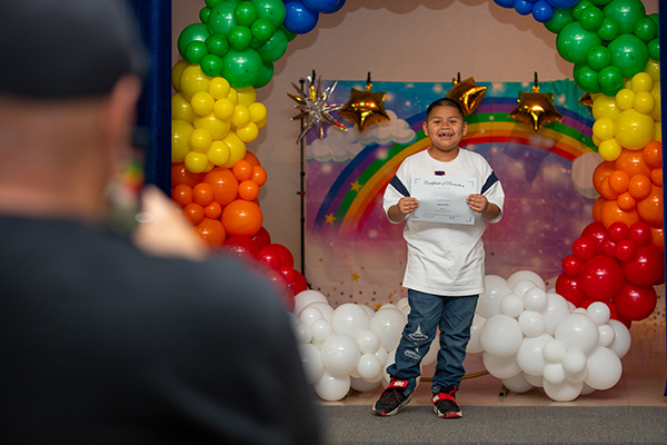 A dad takes a photo of his young son holding up a promotion certificate under a rainbow balloon arch