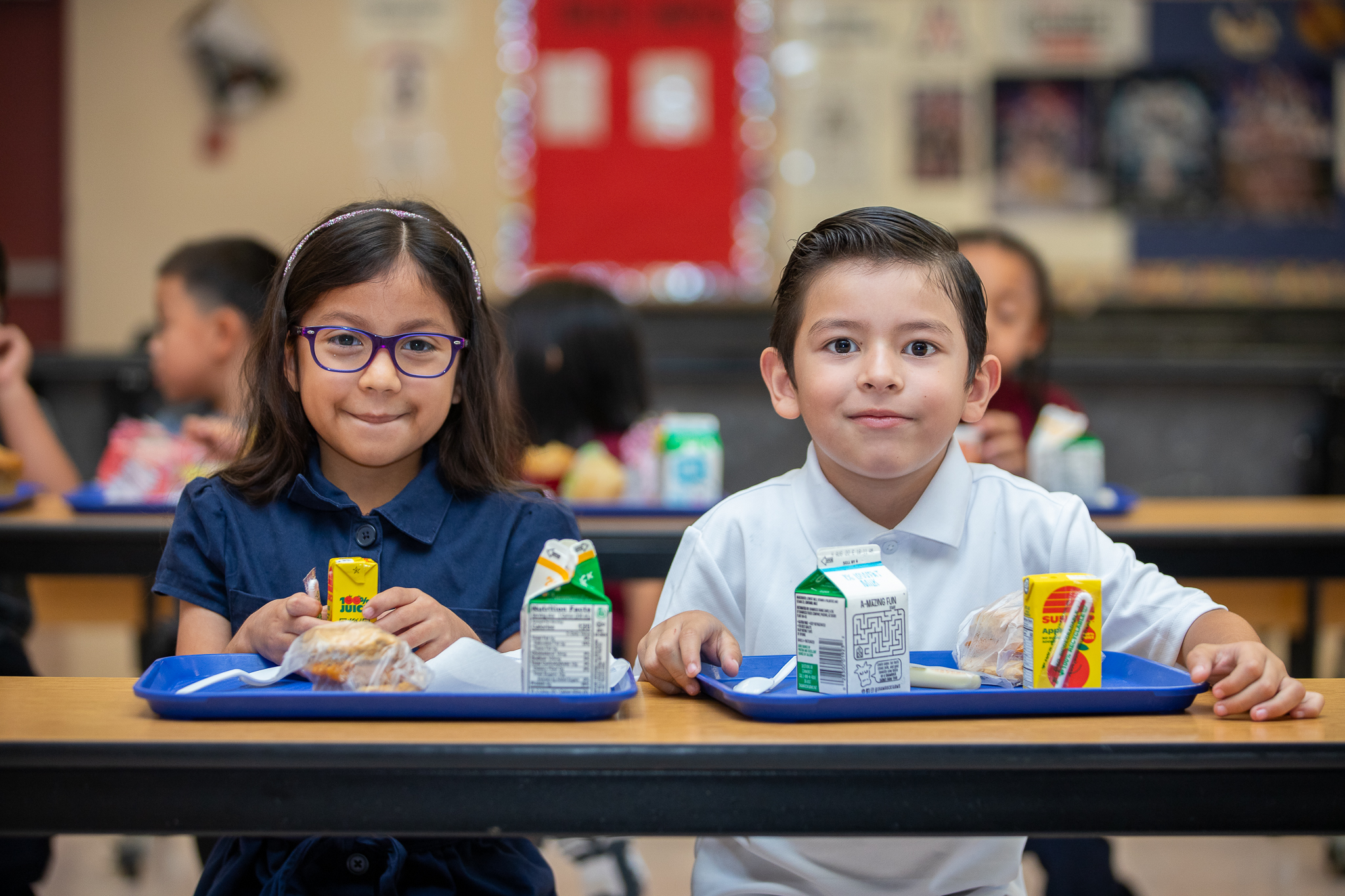 A girl and boy smile with their breakfasts in the cafeteria